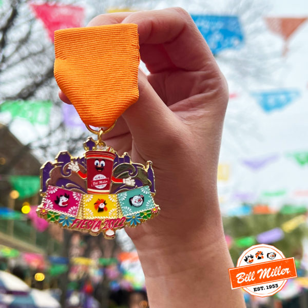 Hand holding Bill Miller Fiesta medal up in front of hanging papel picados. Fiesta medal with mascot Sweetie and cow, chicken, and pig papel picado with the words "Fiesta 2022" surrounded by flowers.