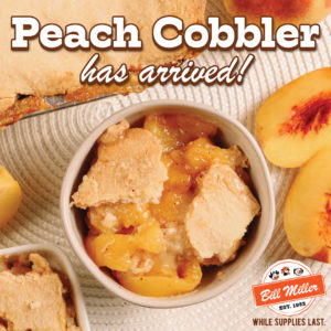 Peach cobbler has arrived! While supplies last. Image of a bowl of peach cobbler on a beige placemat with a pan of peach cobbler peeking in with fresh peaches placed beside the bowl and pan.