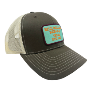 Image includes a dark tan trucker hat with a teal patch outlined with purple stitching. The patch reads, “Bill Miller Bar-B-Q Texas Est. 1953” in orange stitching.
